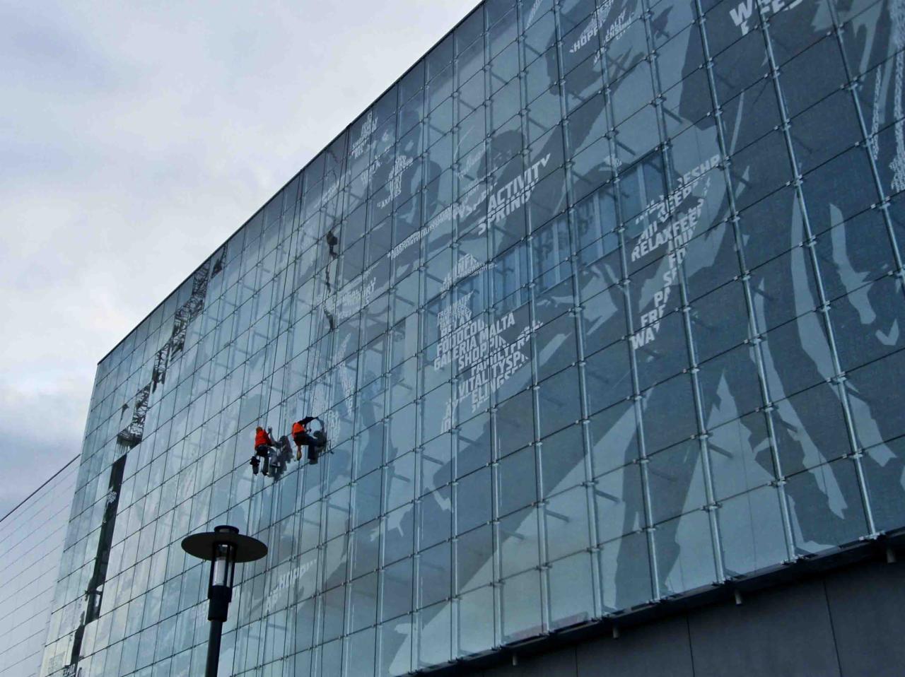 Dwóch alpinistów w pomarańczowych kombinezonach czyści szklaną fasadę budynku z nadrukowanymi napisami, widoczne liny i platformy robocze.