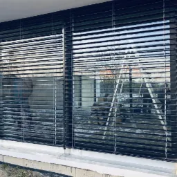 Zamknięte, grafitowe żaluzje poziome w oknie, z widoczną drabiną i elementami budowy na zewnątrz.