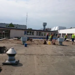 Ekipa budowlana w żółtych kamizelkach i kaskach usuwa gruz i demontuje elementy dachu na płaskim dachu budynku przemysłowego, w tle widoczne inne budynki i zieleń, na dachu leżą płyty betonowe...