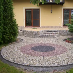 Okrągły wzór z kostki granitowej w odcieniach szarości, różu i beżu na podjeździe przed domem z kamiennymi schodami.