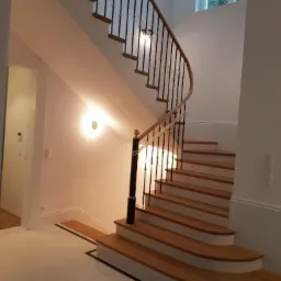 Wnętrze domu z eleganckimi drewnianymi schodami z czarną balustradą i złotymi detalami, oświetlonymi przez okrągłe lampy ścienne, widoczne białe ściany i podłoga z czarnym akcentem.