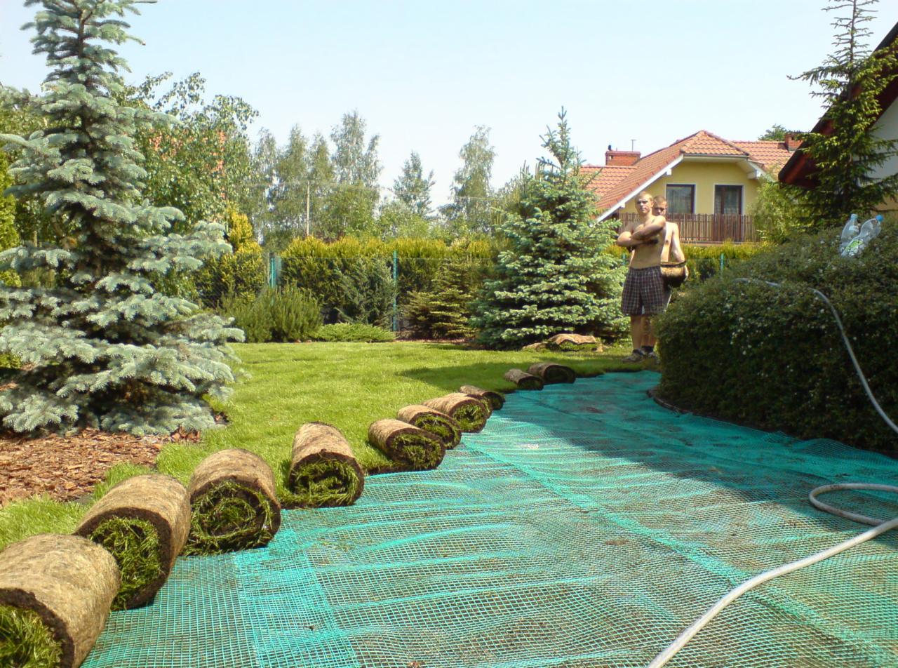 Rozwinięta siatka i rolki trawy w ogrodzie, przygotowane do ułożenia, w tle dom z czerwonym dachem i dwie osoby.