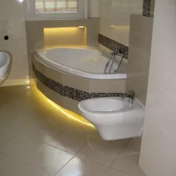 Nowoczesna łazienka z narożną wanną podświetloną LED i bidetem, wykończona beżowymi płytkami z mozaikowym akcentem.