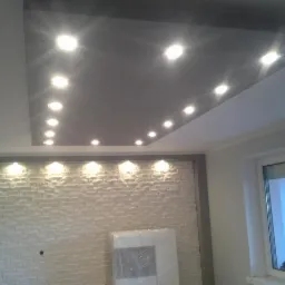 Szary sufit podwieszany z oświetleniem punktowym, biała ściana z dekoracyjną fakturą cegły i okno. Widok na wykończone wnętrze z bliska.