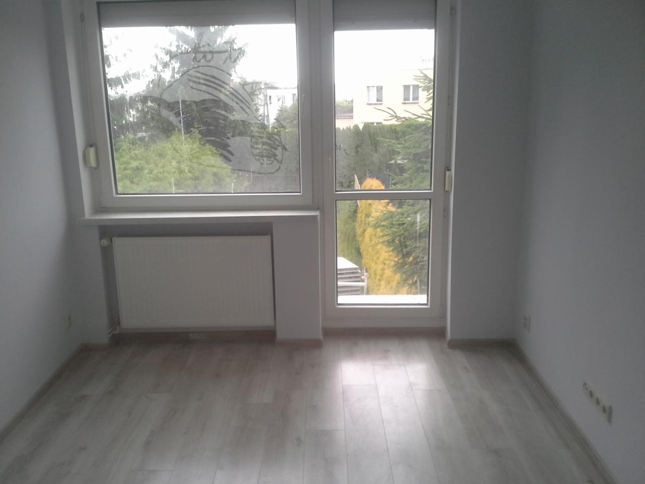 Pusty pokój z panelami podłogowymi w odcieniach szarości, białe ściany, okno z widokiem na zieleń i grzejnik. Pokój przygotowany do wykończenia.