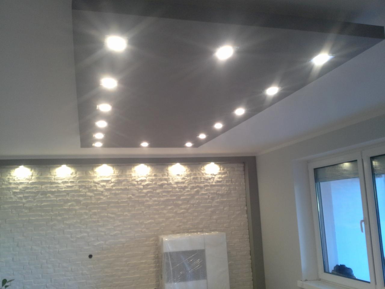 Szary sufit podwieszany z oświetleniem punktowym, biała ściana z dekoracyjną fakturą cegły i okno. Widok na wykończone wnętrze z bliska.