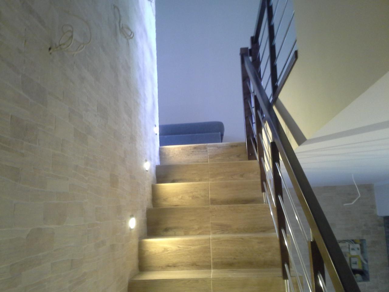 Klatka schodowa z drewnopodobnymi płytkami i oświetleniem punktowym w ścianie. Balustrada z ciemnego drewna i metalu. Widok z dołu schodów.