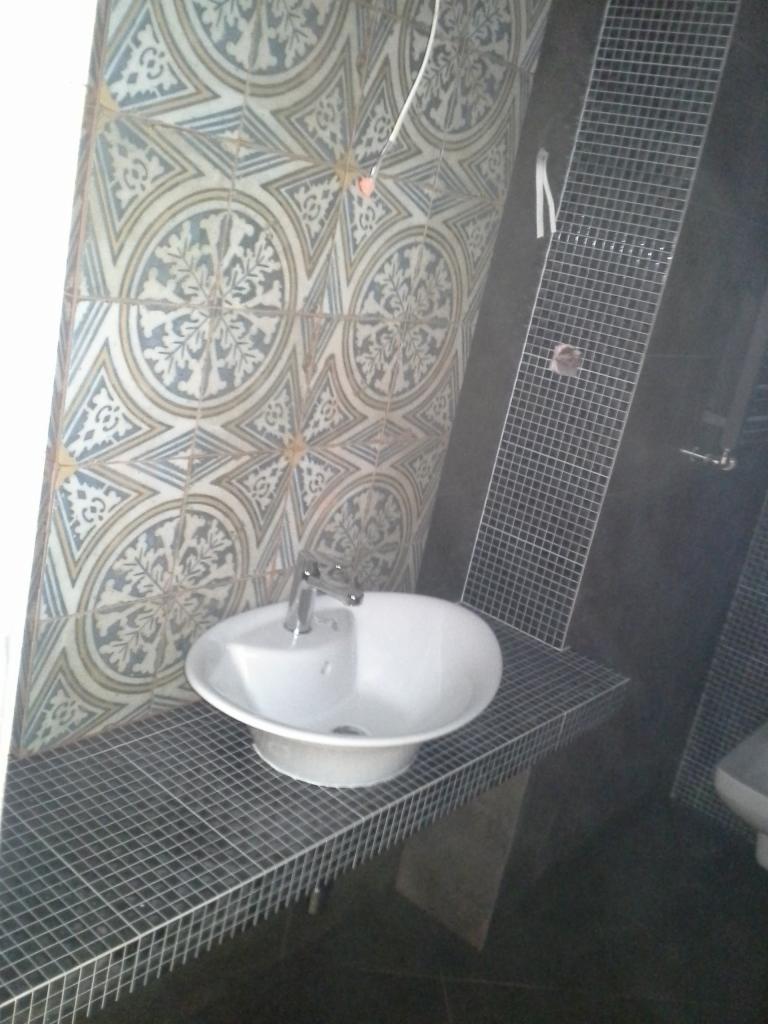 Wykończona łazienka: umywalka z baterią na blacie z mozaiki, ściany wyłożone płytkami z wzorem i szarą mozaiką. Widok na fragment toalety.