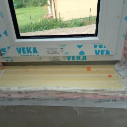 Montaż okna z pianką uszczelniającą i taśmą izolacyjną marki VEKA, widok na zewnątrz budynku z zielenią.