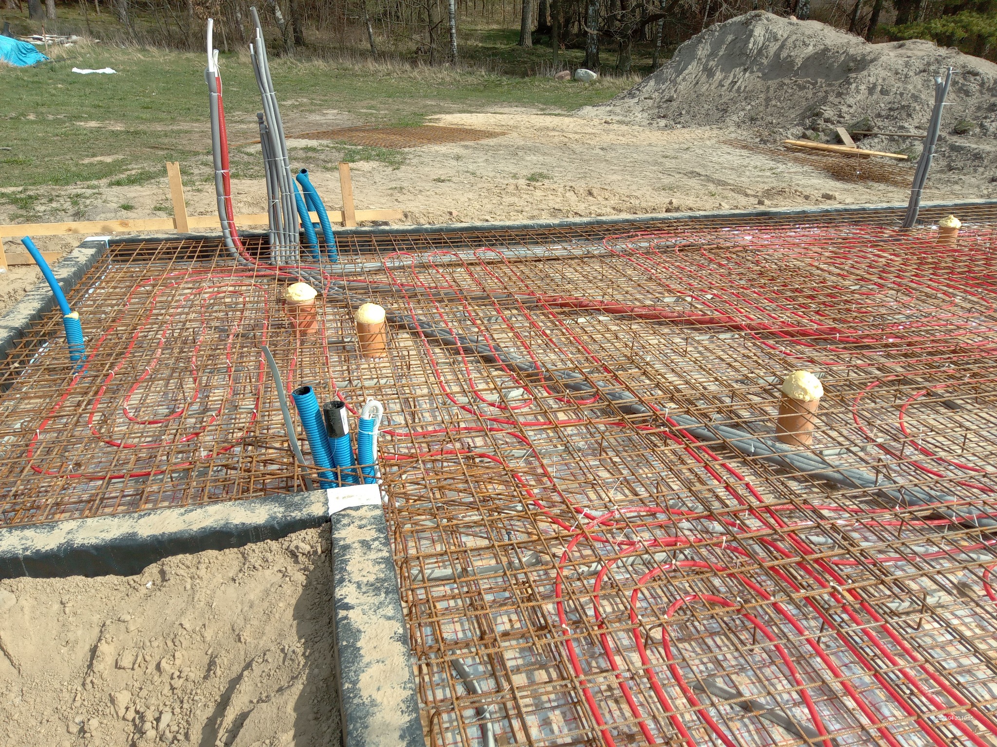 Przygotowanie do wylewki fundamentowej z instalacją ogrzewania podłogowego, rurki grzewcze w kolorze czerwonym ułożone na siatce zbrojeniowej, wystające rury instalacyjne zabezpieczone pianką...