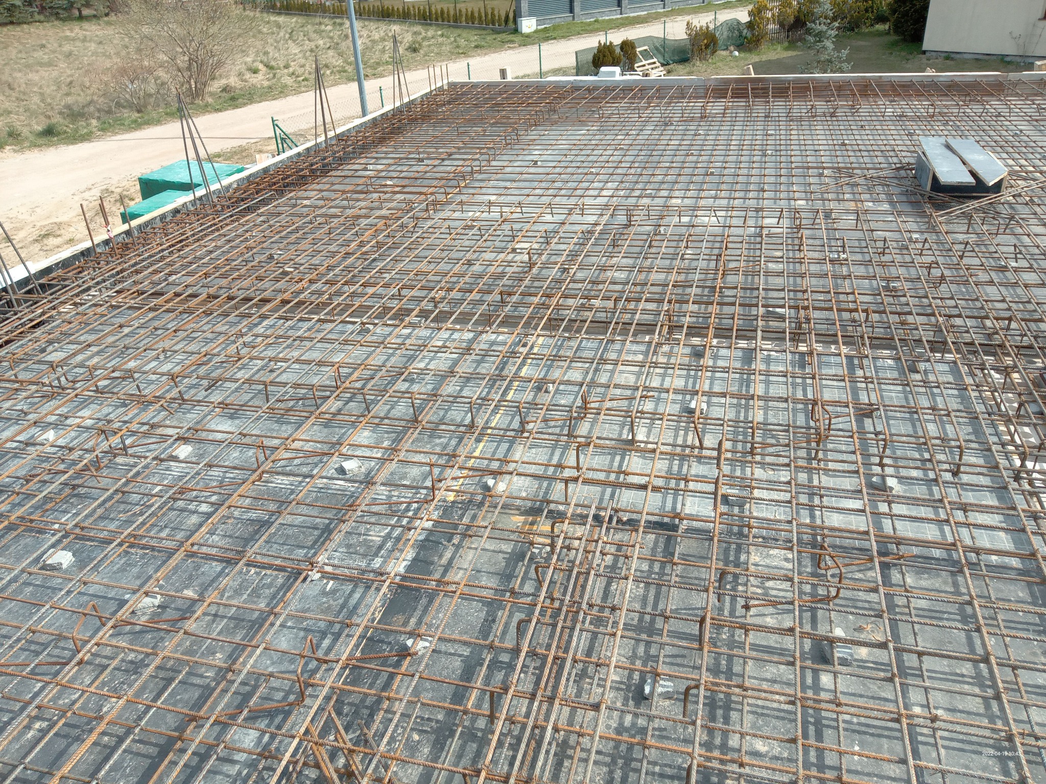 Siatka zbrojeniowa ułożona na betonowym fundamencie, widok z góry, przygotowana do zalania betonem, z widocznymi elementami dystansowymi i fragmentami otoczenia.