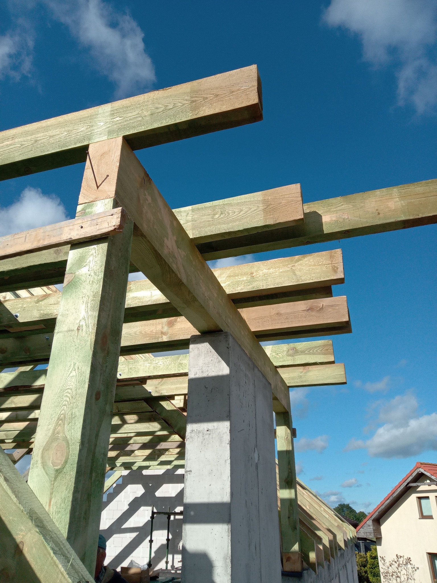 Konstrukcja drewniana dachu w trakcie budowy, widoczne impregnowane belki, betonowy filar, niebo z chmurami w tle.