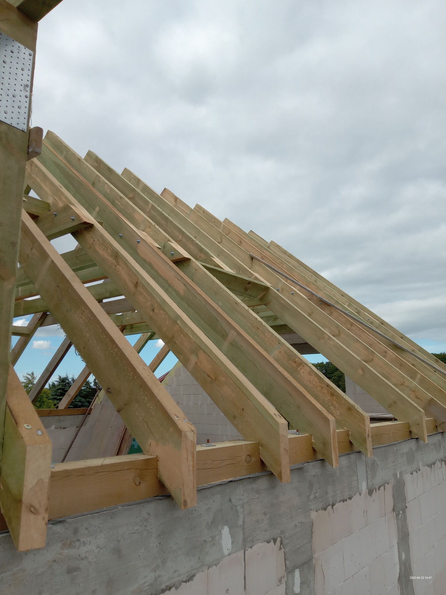 Konstrukcja drewniana dachu z widocznymi krokwiami mocowanymi na metalowe płytki perforowane, w tle pochmurne niebo.