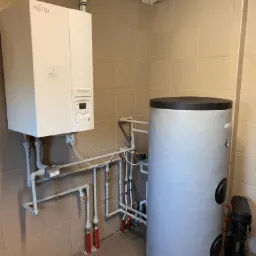 Biały, wiszący piec gazowy Fujitsu Waterstage z szarym zasobnikiem wody, połączone rurami w pomieszczeniu z beżowymi płytkami.
