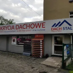 Fasada sklepu z pokryciami dachowymi, szyld z napisem 'POKRYCIA DACHOWE' i logo 'DACH STAL', ekspozycja dachówek i blach trapezowych przed wejściem, pochmurne niebo w tle.