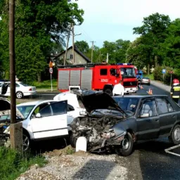 Miejsce wypadku drogowego z uszkodzonymi samochodami, w tle wóz strażacki i policjant kierujący ruchem.