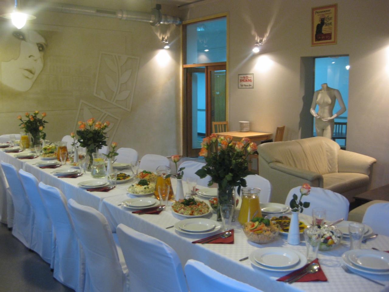 Długi stół nakryty białym obrusem i krzesła z białymi pokrowcami, przygotowany do cateringu, z aranżacją kwiatową z róż, szklanymi naczyniami z sokiem i sałatkami, w tle dekoracje ścienne, sofa...