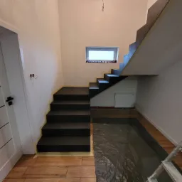 Wnętrze w trakcie malowania: ciemne schody zabezpieczone taśmą malarską, podłoga przykryta folią ochronną, widoczne kable elektryczne wystające ze ściany.