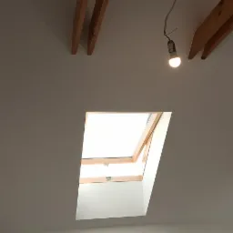 Poddasze po tynkowaniu z widocznymi drewnianymi belkami, oknem dachowym i prowizorycznym oświetleniem żarówką.