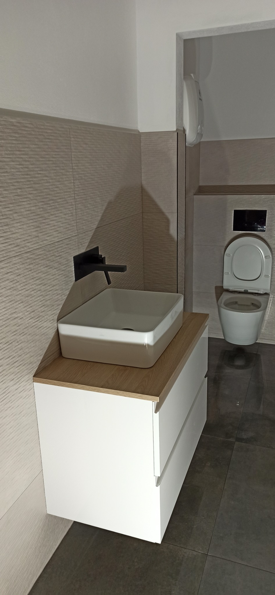 Nowoczesna łazienka z umywalką nablatową w kolorze beżowo-białym, szafką z białymi frontami i blatem w odcieniu drewna, czarną armaturą i podwieszaną toaletą.