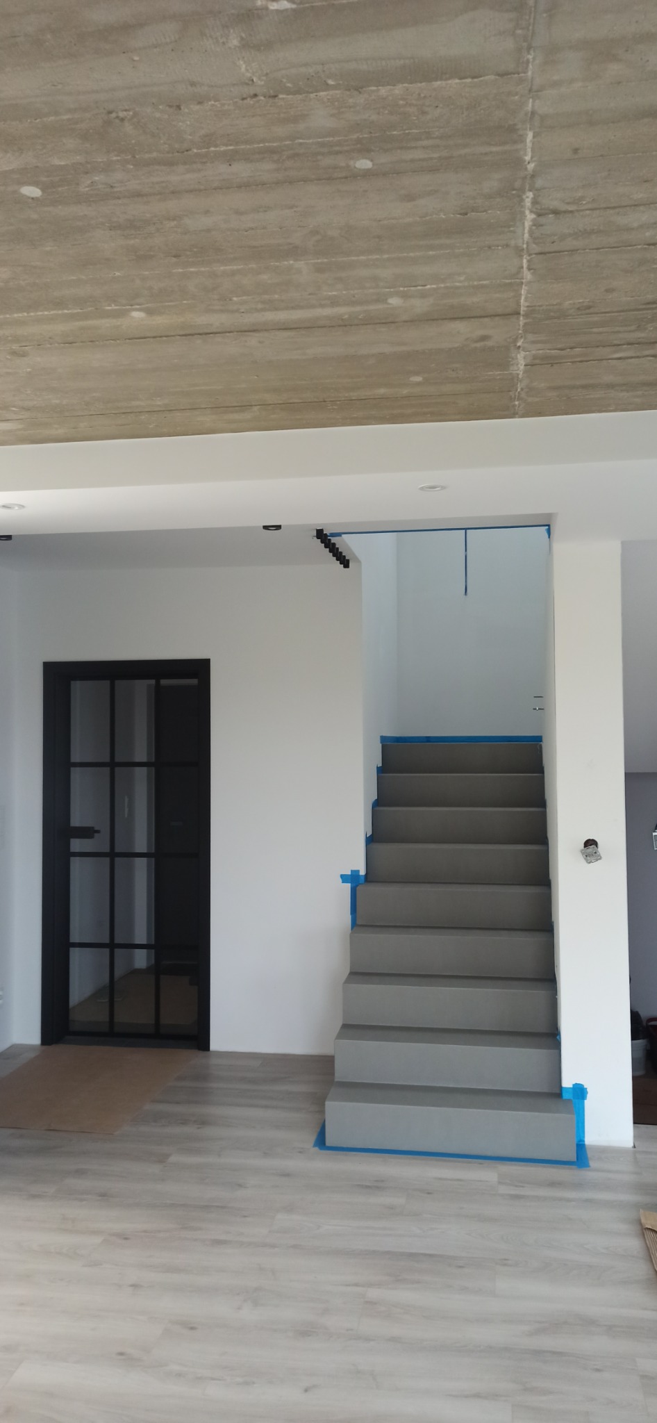 Wnętrze w trakcie prac wykończeniowych: surowy betonowy sufit, pomalowane na biało ściany, szara klatka schodowa zabezpieczona niebieską folią malarską, czarne drzwi z szybami, jasna podłoga.