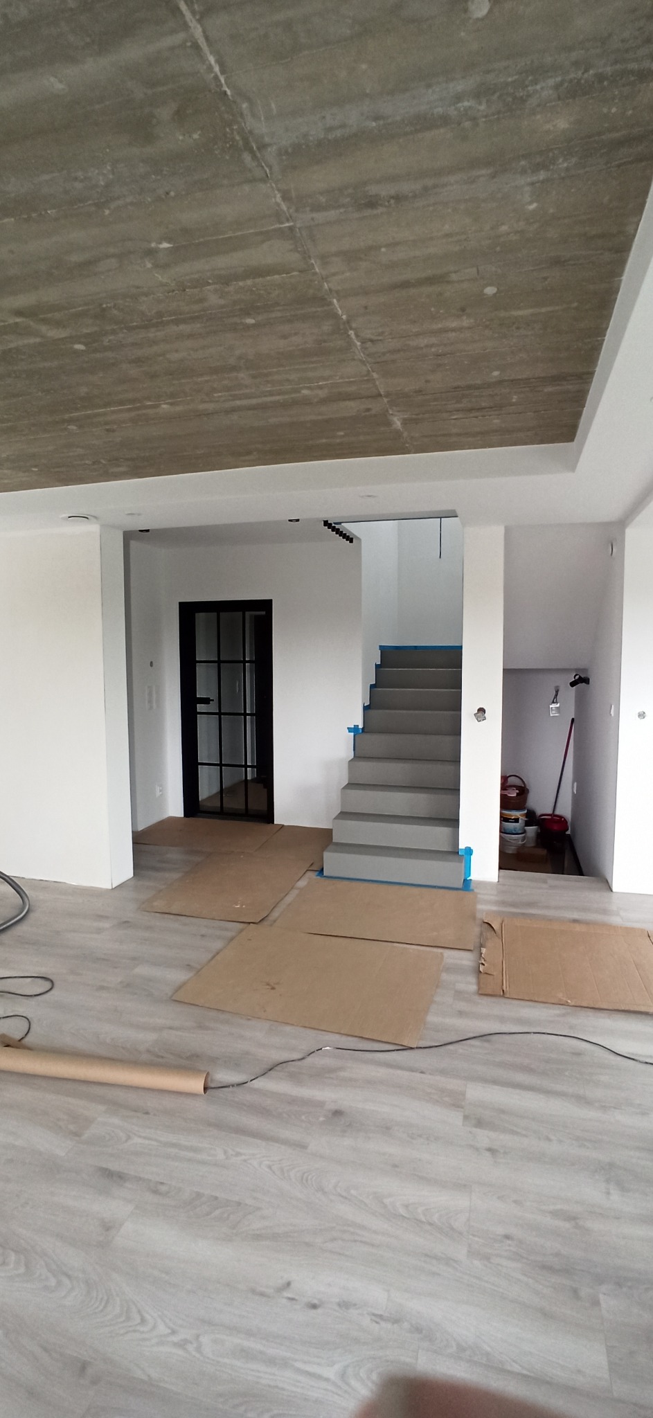 Wnętrze w trakcie prac wykończeniowych: surowy betonowy sufit, świeżo pomalowane białe ściany, szare schody zabezpieczone niebieską taśmą malarską i podłoga częściowo zakryta kartonem ochronnym.