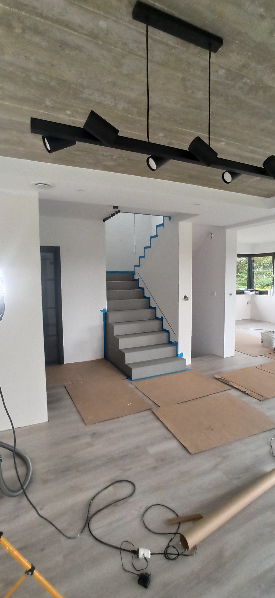 Wnętrze domu w trakcie prac wykończeniowych: zabezpieczone schody z pomalowanymi stopniami, karton na podłodze, nowoczesne oświetlenie sufitowe, świeżo pomalowane ściany.