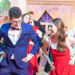 Grupa młodych ludzi tańczy na parkiecie podczas uroczystości, mężczyzna w niebieskim garniturze i muszce, kobieta w czerwonej sukience śmieje się, w tle dekoracje świetlne.