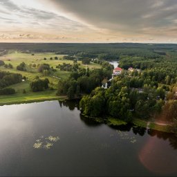 Studio Foto Video Michał Słupczewski - Szeroki kadr z drona ukazuje jezioro otoczone lasami i polami w promieniach zachodzącego słońca. W tle widoczny budynek z czerwonym dachem.