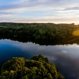 Studio Foto Video Michał Słupczewski - Spokojne jezioro otoczone lasem, uchwycone z lotu ptaka o zmierzchu. Woda odbija niebo, tworząc malowniczy krajobraz. Promienie zachodzącego słońca dodają ciepła.