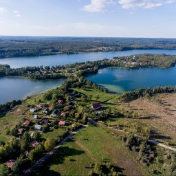 Studio Foto Video Michał Słupczewski - Panoramiczny widok z lotu ptaka na malownicze jezioro z wyspami i zabudowaniami, otoczone lasami i polami w słoneczny dzień. Ujęcie z góry ukazuje detale krajobrazu.