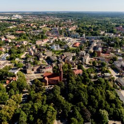 Studio Foto Video Michał Słupczewski - Panorama miasta z lotu ptaka w słoneczny dzień. Widoczne budynki, zieleń drzew i parków, kościół z czerwoną dachówką. Ujęcie z góry, perspektywa szerokokątna.