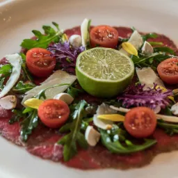Carpaccio wołowe z rukolą, pomidorkami cherry, plasterkami parmezanu, grzybami i limonką na białym talerzu, oświetlone ciepłym światłem.