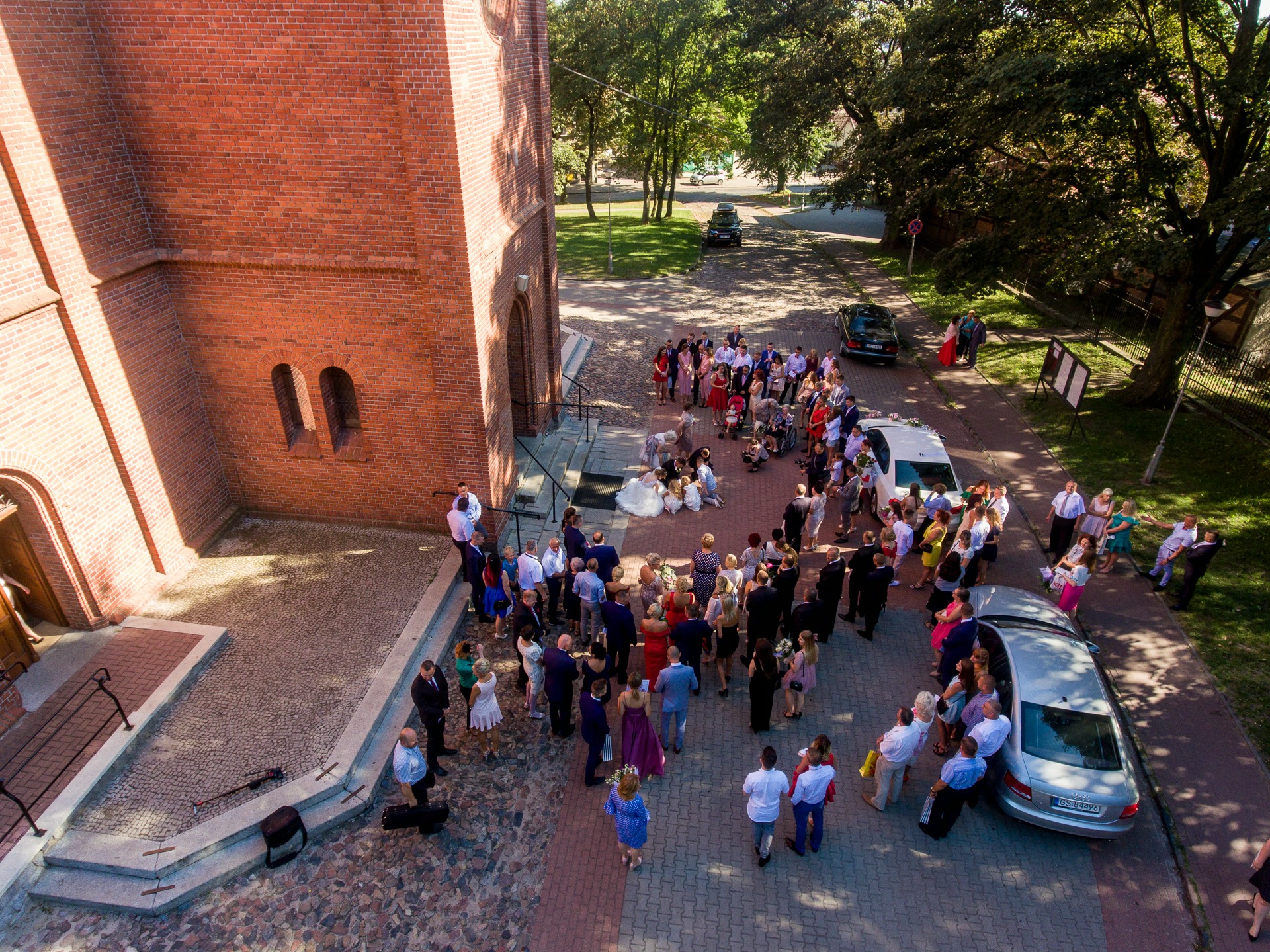 Zdjęcie z drona ukazuje grupę gości weselnych przed ceglanym kościołem, z parą młodą na schodach i samochodami w tle, w słoneczny dzień.