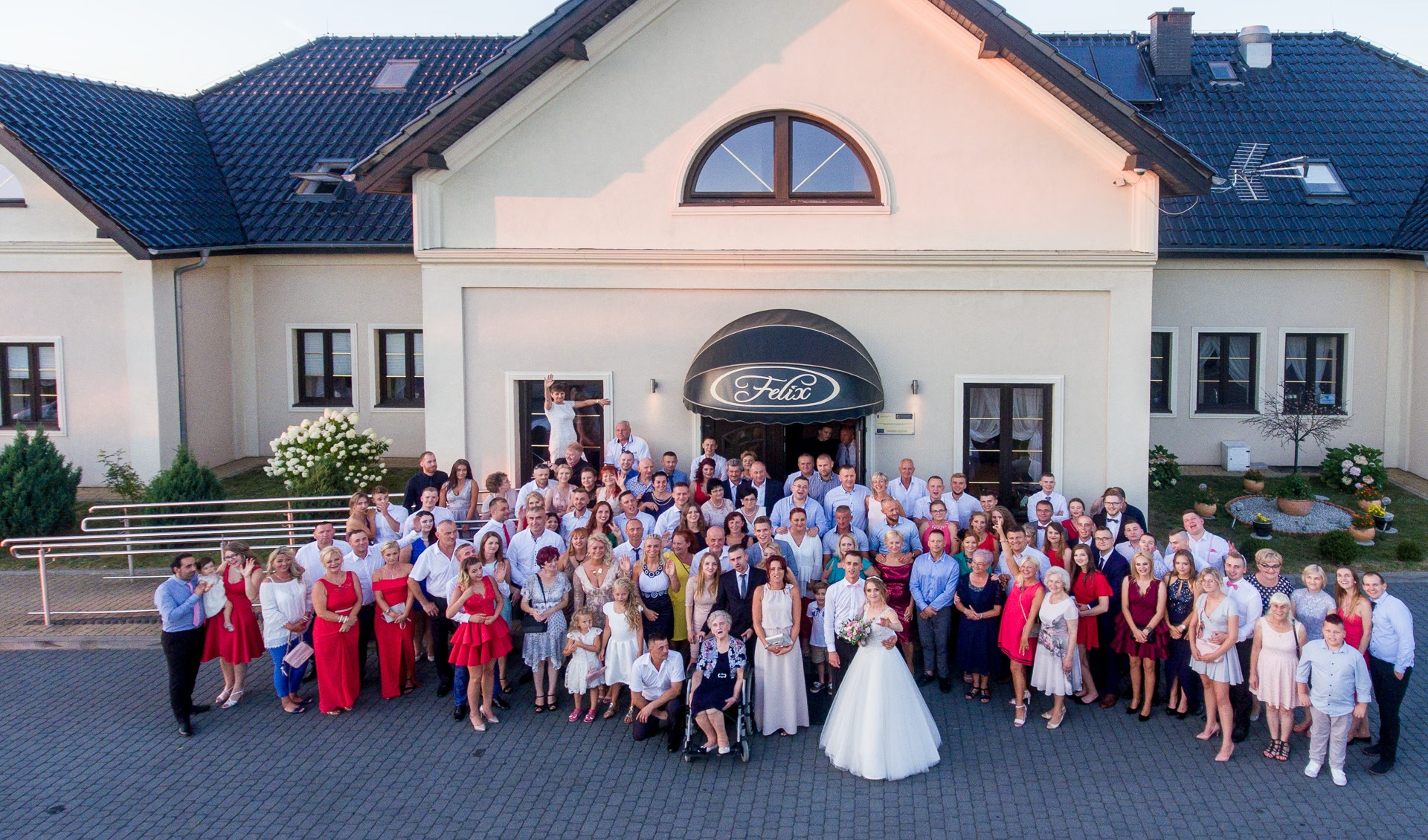 Grupowe zdjęcie ślubne przed budynkiem z markizą 'Felix', panna młoda w białej sukni na pierwszym planie, goście weselni w eleganckich strojach, ciepłe światło.