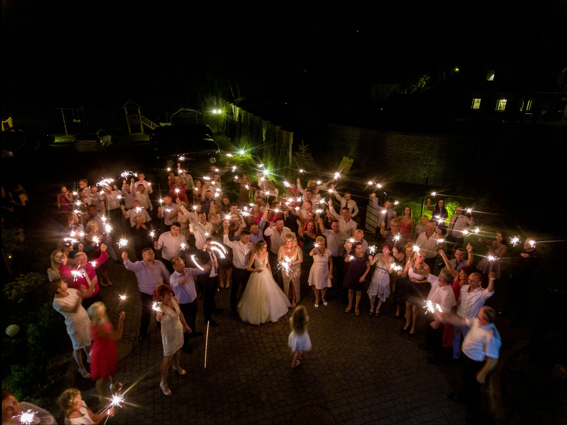 Nocne ujęcie z góry: para młoda otoczona gośćmi weselnymi trzymającymi zapalone zimne ognie, tworząc iskrzący krąg światła na tle ciemnego otoczenia.