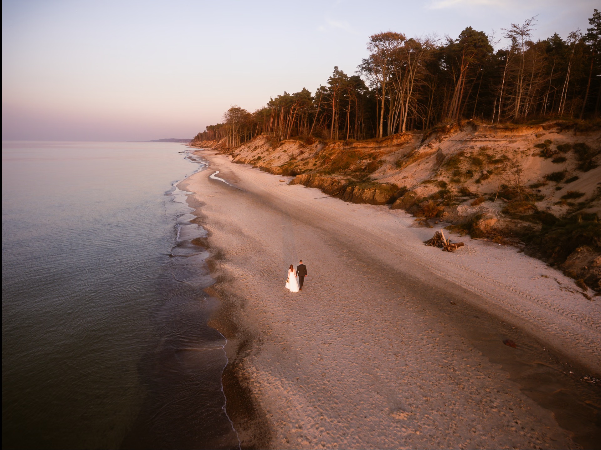 Para młoda spaceruje brzegiem morza o zachodzie słońca. Widok z góry ukazuje piaszczystą plażę, las na klifie i spokojną wodę. Romantyczna sceneria.