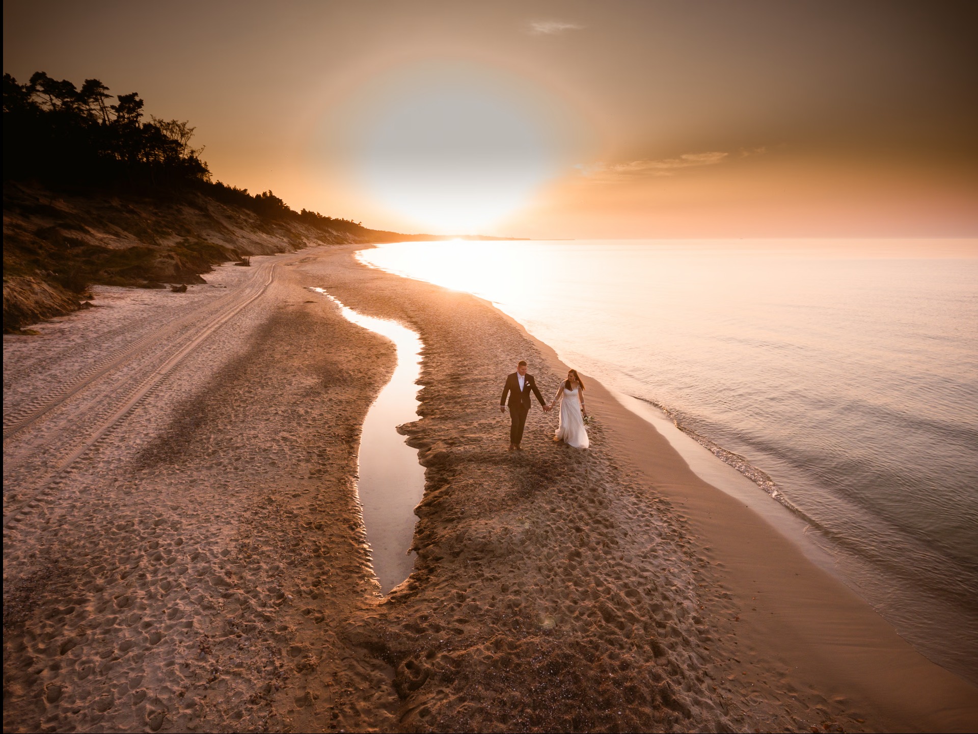 Para młoda spaceruje brzegiem morza o zachodzie słońca. Panna młoda w białej sukni, pan młody w garniturze. Romantyczna sceneria, ciepłe światło.