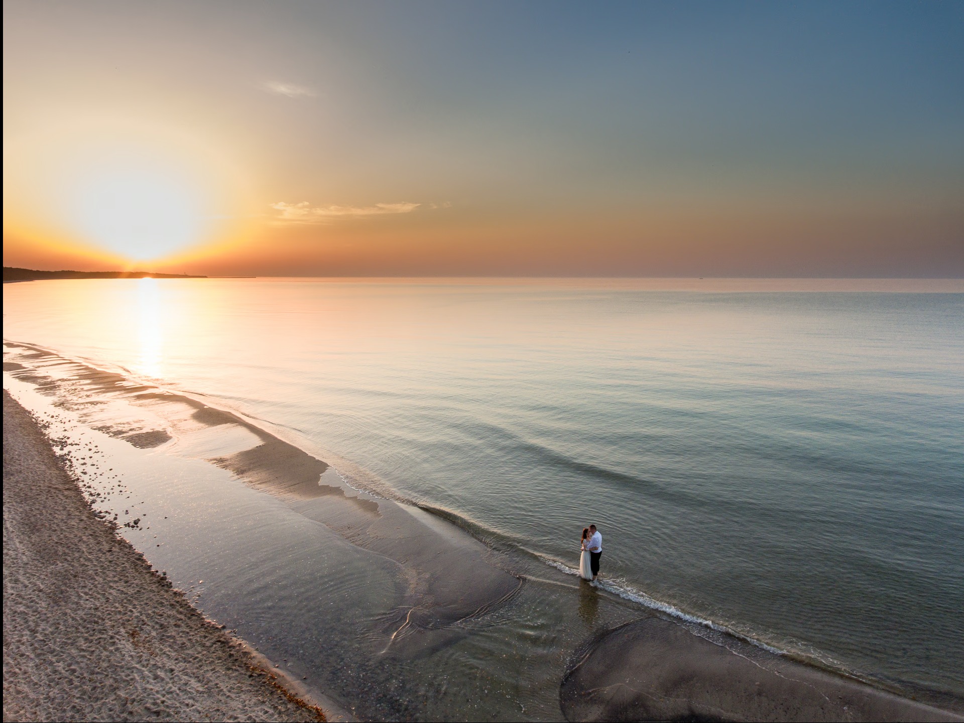 Para młoda w objęciach na brzegu morza o wschodzie słońca. Szeroki kadr z lotu ptaka, spokojna woda, delikatne fale. Romantyczna sesja ślubna na plaży.