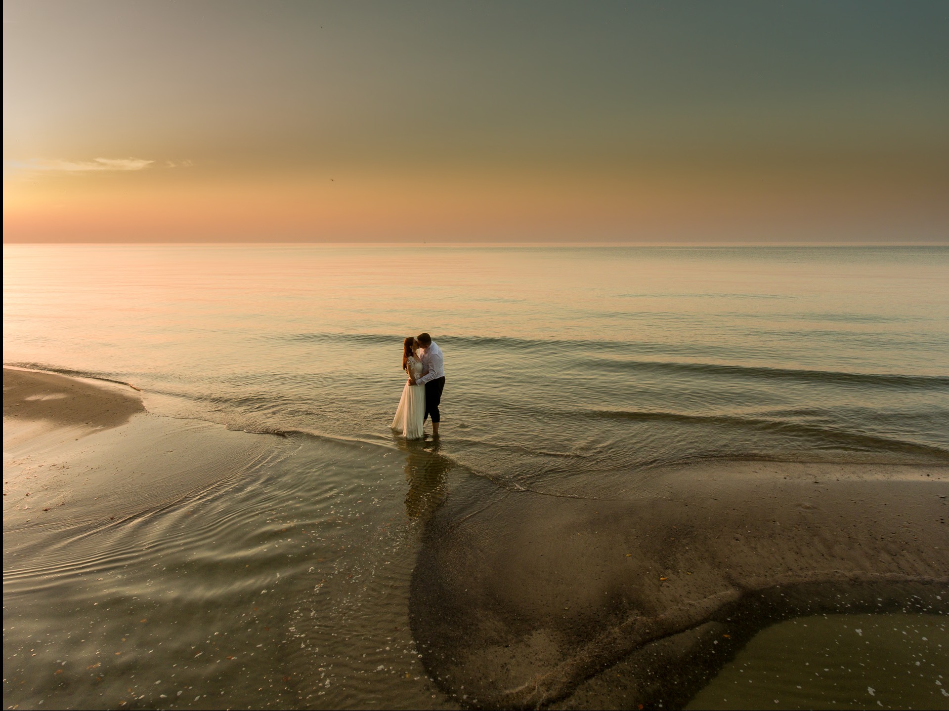 Para młoda w Słupsku całuje się na plaży o zachodzie słońca. Romantyczna sesja zdjęciowa nad morzem, delikatne fale obmywają brzeg. Ciepłe barwy nieba.