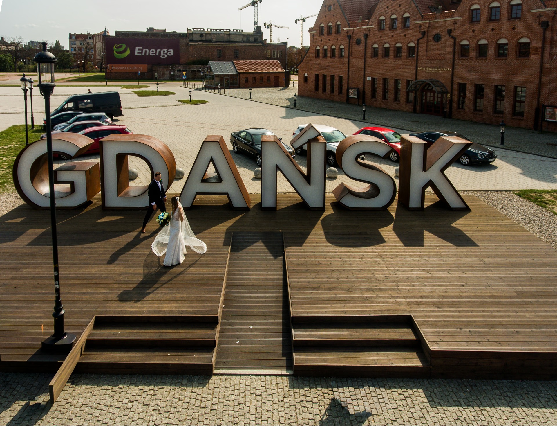 Para młoda w Gdańsku pozuje na drewnianym podeście z napisem 'GDAŃSK' w tle ceglane budynki i samochody. Ujęcie z góry, oświetlenie naturalne.