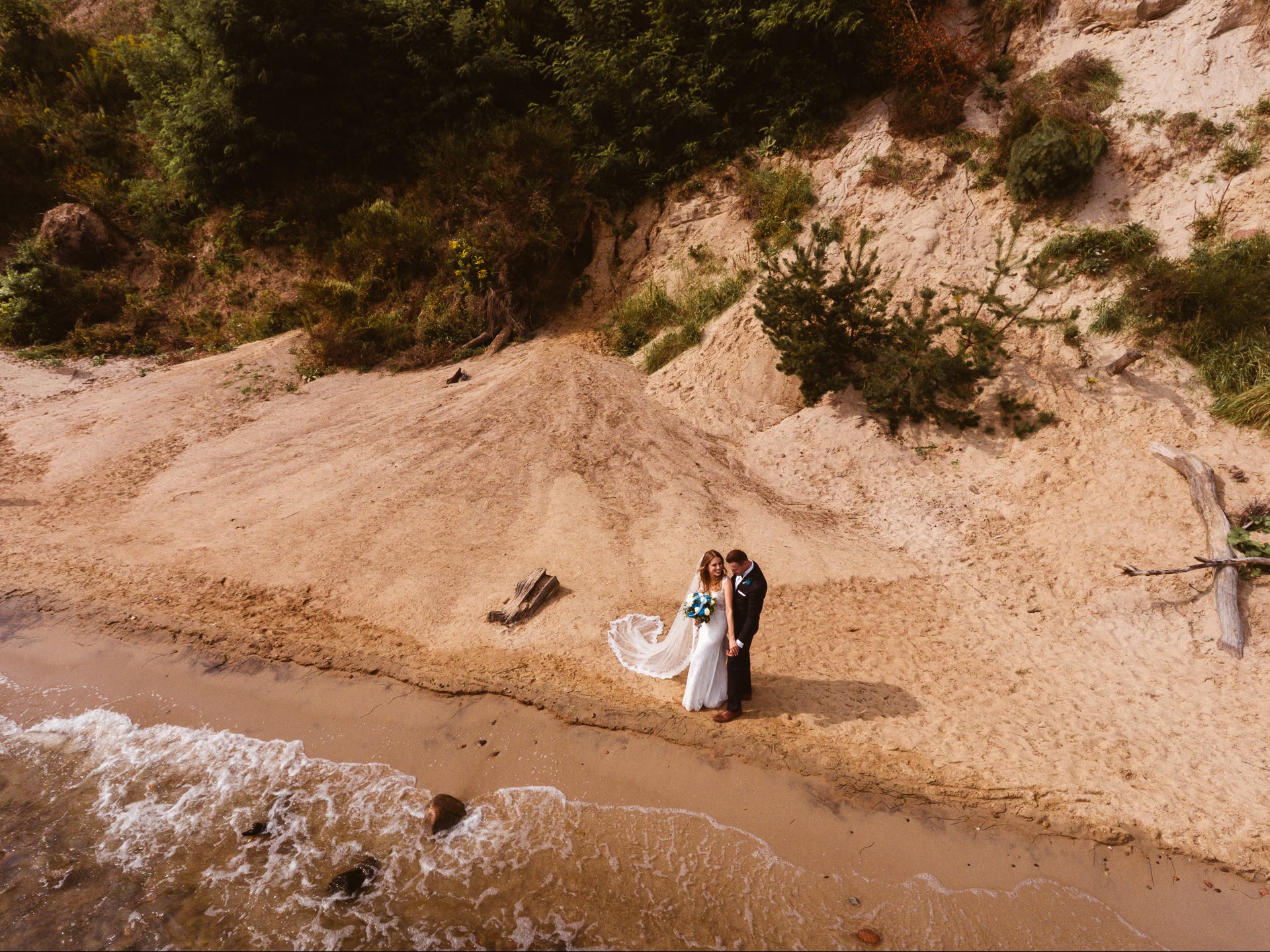 Para młoda na piaszczystej plaży z klifem w tle, ujęcie z góry. Panna młoda w białej sukni z bukietem, pan młody w ciemnym garniturze. Romantyczna sceneria.
