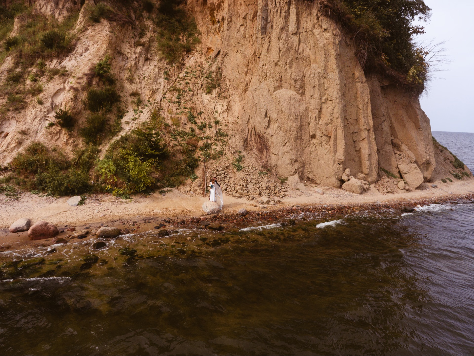 Para młoda w białych strojach stoi na kamieniu na plaży u stóp klifu porośniętego zielenią, z widokiem na morze. Ujęcie z lotu ptaka.