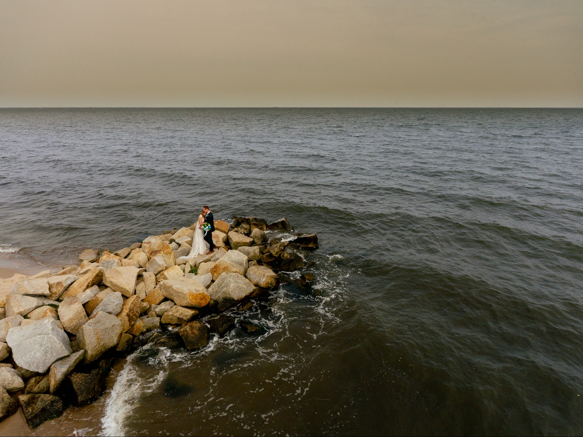 Para młoda na kamiennym falochronie, szeroki kadr z lotu ptaka. Morze i niebo w stonowanych barwach. Panna młoda w białej sukni, pan młody w ciemnym garniturze.
