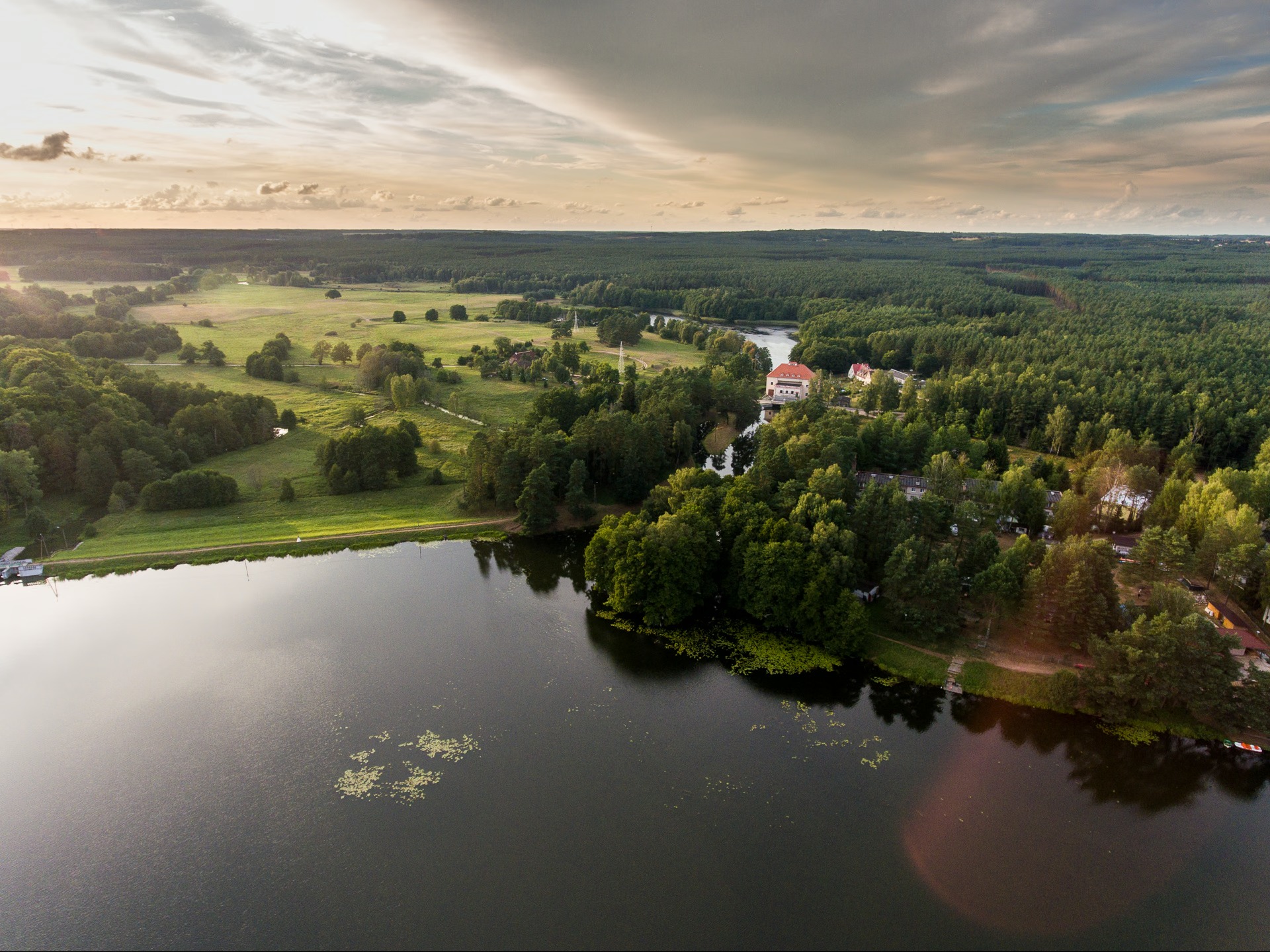 Szeroki kadr z drona ukazuje jezioro otoczone lasami i polami w promieniach zachodzącego słońca. W tle widoczny budynek z czerwonym dachem.