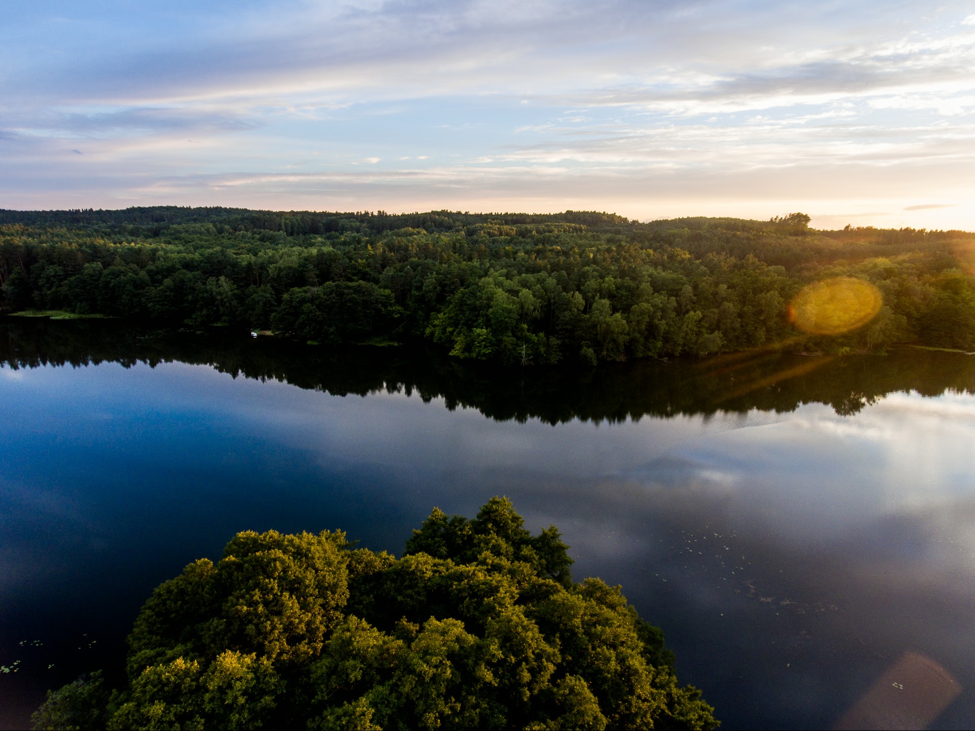 Spokojne jezioro otoczone lasem, uchwycone z lotu ptaka o zmierzchu. Woda odbija niebo, tworząc malowniczy krajobraz. Promienie zachodzącego słońca dodają ciepła.