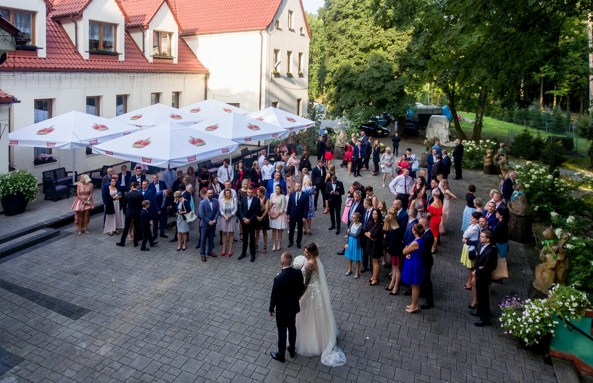Górny kadr na parę młodą i gości weselnych na dziedzińcu z parasolami Tyskie, w tle budynek z czerwonym dachem i zielone drzewa. Ujęcie z drona.