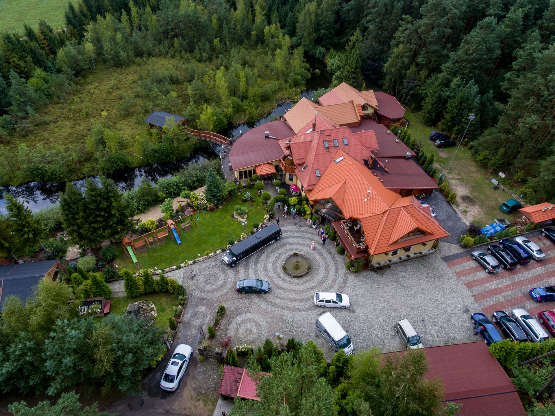 Szeroki kadr z lotu ptaka ukazuje kompleks budynków z czerwonymi dachami, otoczonych zielenią, z fontanną i zaparkowanymi autami na dziedzińcu. W tle las i mały staw.