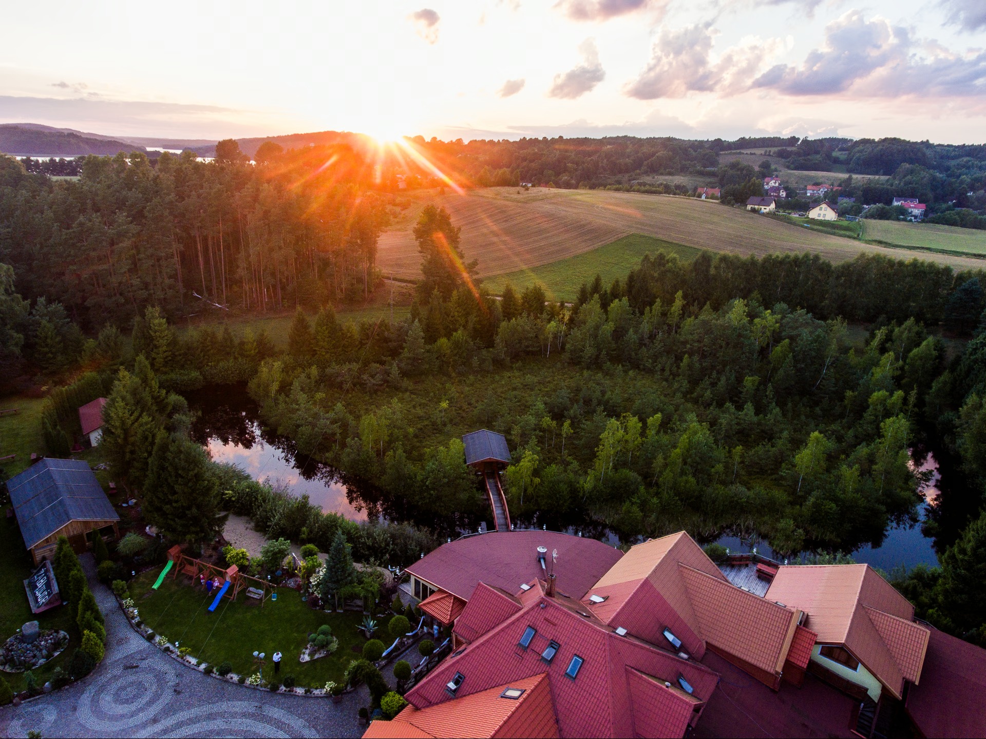 Panoramiczny widok z lotu ptaka na malowniczy krajobraz z jeziorem, lasami, polami i budynkami o czerwonych dachach w promieniach zachodzącego słońca.