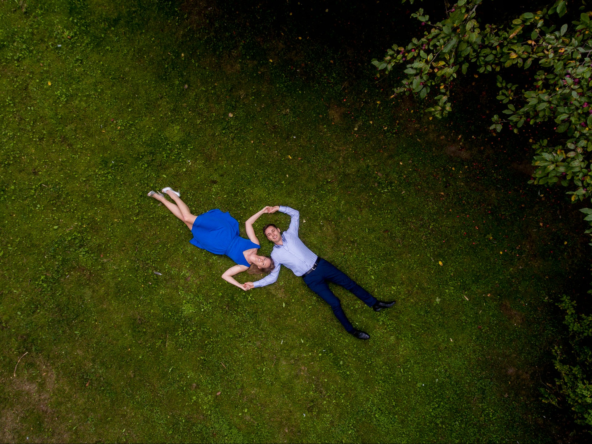 Para leżąca na trawie, widok z góry. Kobieta w niebieskiej sukience, mężczyzna w koszuli i spodniach. Tworzą serce z rąk. Ujęcie z drona, zieleń trawy i drzew.